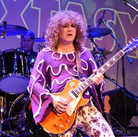 A man in a flamboyant purple top plays an orange guitar. He looks as though he is concentrating.