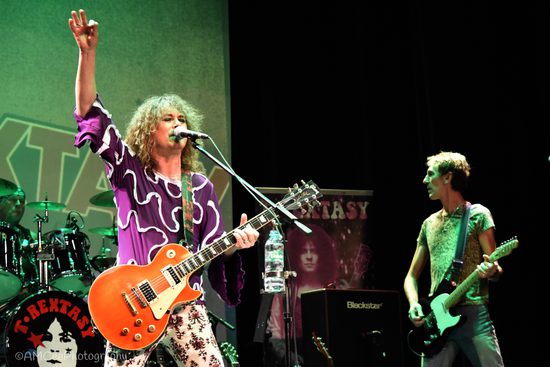 A man in a flamboyant purple top with shoulder length hair plays an orange guitar with one arm in the air. The lighting around him is green.