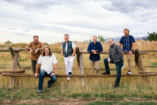 6 band members sit and stand next to an old wooden fence