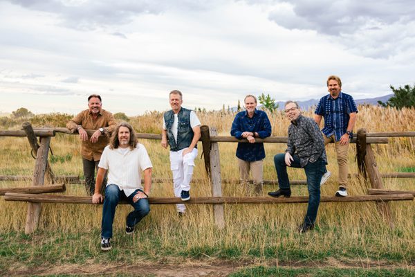 6 band members sit and stand next to an old wooden fence