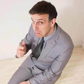 A man in a grey suit sat on the floor, pointing at the camera.