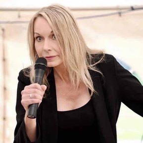 A woman in a black top and jacket holds a microphone and looks alarmed.