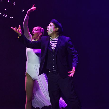 A mime in a stripy top and black jacket throws white confetti in the air. A woman in a white dress stands behind him, amazed.