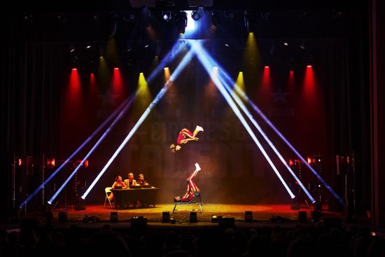 Acrobats flip on the stage. Strobe lighting creates a triangle of white light, with red and yellow lights behind.