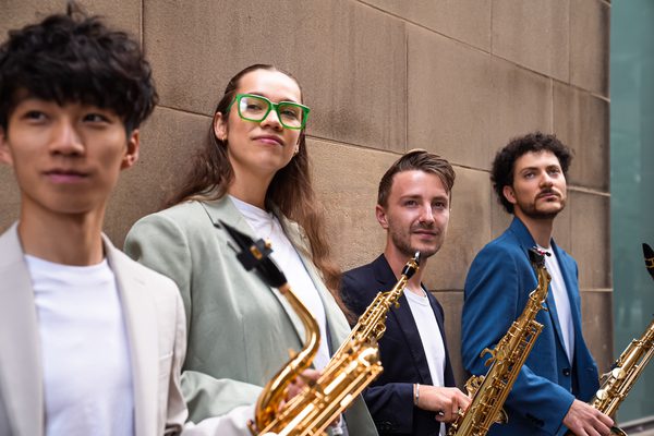 Four saxophonists lean against a wall stone wall. The all wear a white t-shirt and neutral jacket.