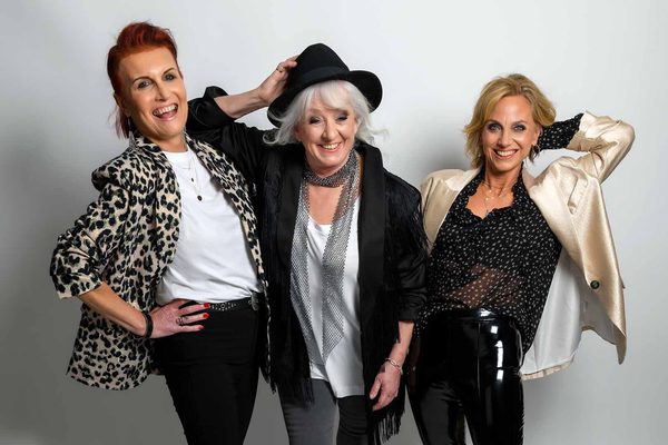 Three older white women laughing. One had red hair and a leopard print jacket, one has grey hair, a black jacket and a black hat, and one has blonde hair and a gold jacket.