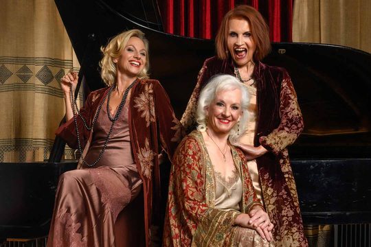 Three women in luxurious gold and red dresses and robes smiling. They are sitting around a grand piano.