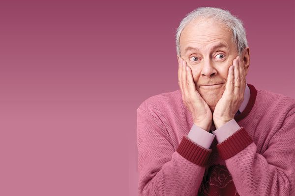 An older man wearing a pink shirt and jumper holds his cheeks in his hands. He is sat against a pink background.