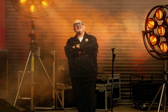 Man with short grey hair and dark rimmed glasses stands back stage. He holds a microphone with his arms crossed smiling towards the ceiling.