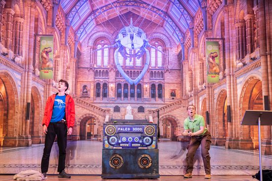 A woman in a green top and a man in a blue top and red jacket stand in front of a backdrop of a large museum.