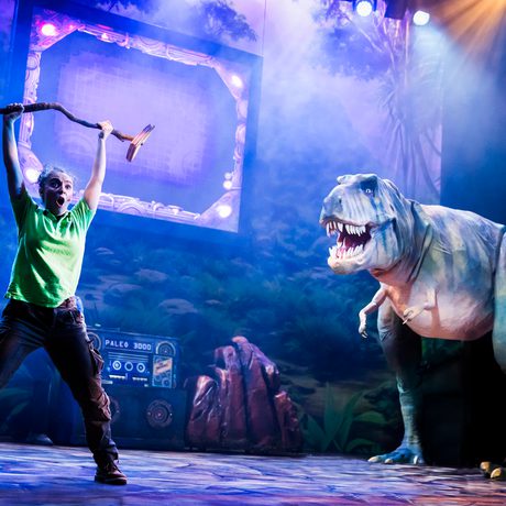 A woman in a green top holding up a large stick at a large T-rex puppet. They are lit in blue.