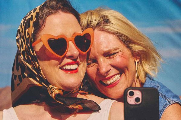 Two women smiling and taking a selfie. One is in a denim top and the other is in a colourful headscarf and orange heart shaped sunglasses.