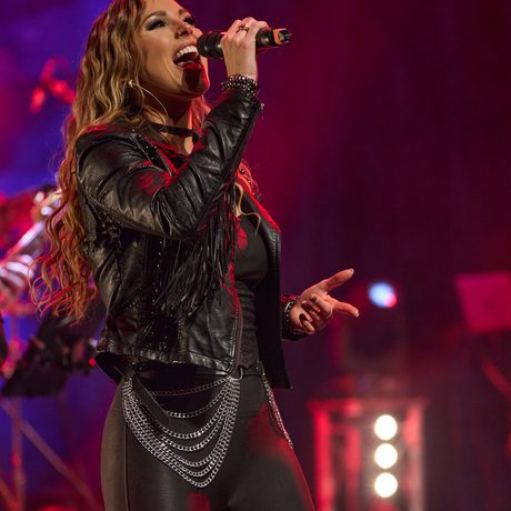 A woman wearing black leather trousers and a black leather jacket sings into a microphone. The stage behind her is lit in blue and red.