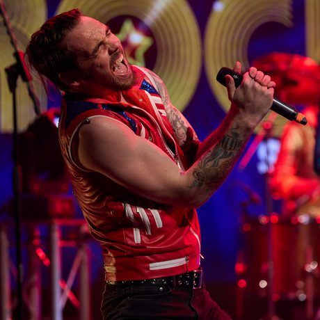 A man in black jeans and a red leather jacket sings loudly into a microphone.