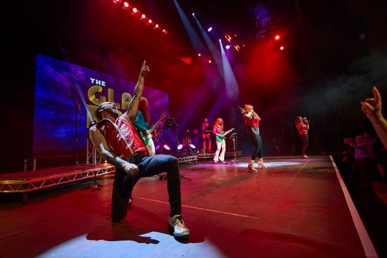 A band playing instruments such as guitar and drums, and three singers, perform on stage to a crowd. A male singer at the front of the image is kneeling down with one hand pointing in the air.