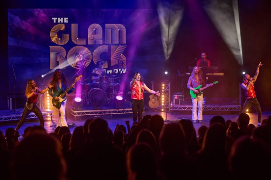 A band playing various instruments including guitar and drums, as well as three singers, perform to a full audience.