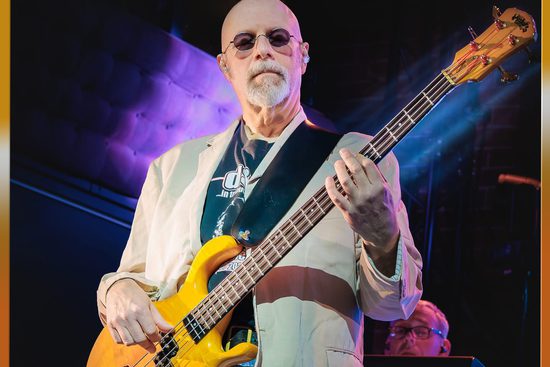 An older, white man with a grey beard wears sunglasses and a cream jacket. He is playing a bright yellow bass guitar.