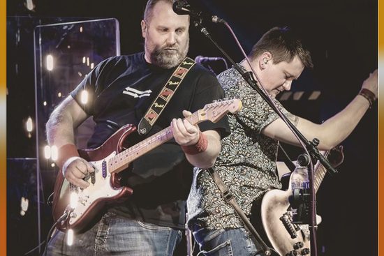 Two white, brunette man play guitars while standing ack-to-back. One is wearing a black t-shirt and the other is wearing a green floral shirt.
