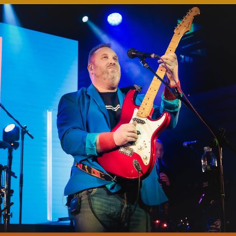 A white, brunette man wearing a bright blue jacket plays a red and white electric guitar.