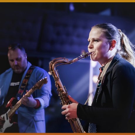 A white, blonde woman wearing a black jacket plays the saxophone.