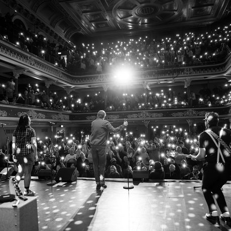 Black and white image of a band playing on a stage. The image is taken from behind the band, so you can see the audience holding up lights.