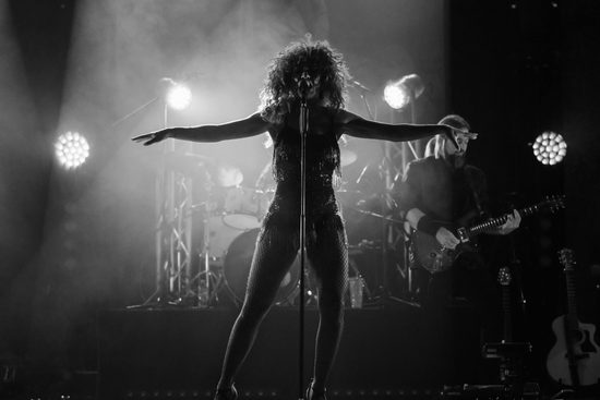 Black and white image of the silhouette of a woman standing on a stage singing. Lights shine from behind her.