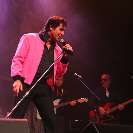 A man in a black shirt and trousers and a bright pink jacket sings into a microphone.