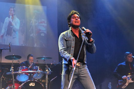 A man in a black shirt, blue jeans and a denim jacket sings with a drummer and guitarist behind him.