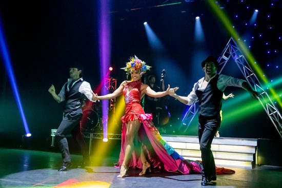 A woman in a bright pink dress, gold high heels, and a flower headdress walks on a stage with a man in a white shirt and black waistcoat on either side of her.