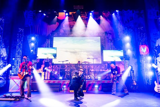 A band perform on a stage with a screen at the pack showing an image of the clouds. The lead singer is crouched down to the ground.