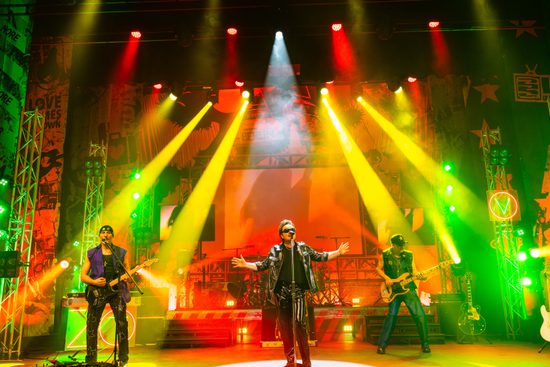 A band wearing leather jackets stand on a stage and perform. The lighting is red, yellow, and green.