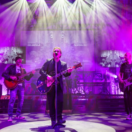 A band wearing black stand on a stage and perform. The lighting is pink and purple.