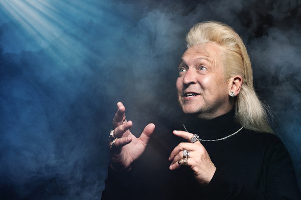 A man in a black polo neck with long blonde hair stands against a blue background. He is pointing and looking up at something, and smiling.
