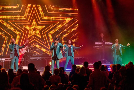 Four black men in green suits singing to an audience. On the screen behind them is a large gold star.