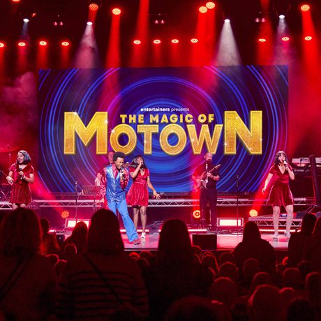 Various performers on stage singing and playing instruments. Three women are wearing red dresses and one man is wearing a blue suit. They are lit with red lighting.