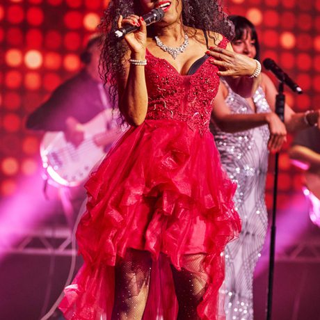 A black woman with long brown curly hair in a red frilly dress that is shorter at the front and longer at the back sings into a microphone.