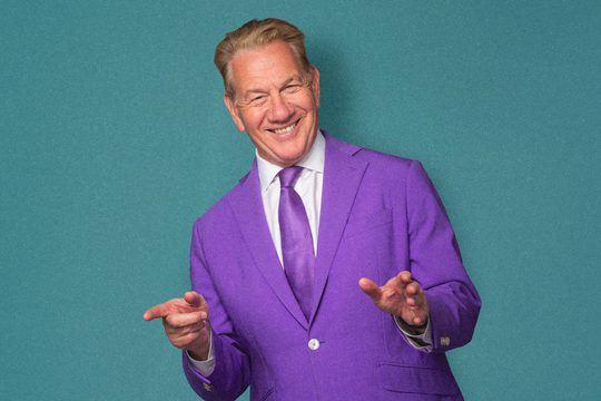 A man in a bright purple suit smiles and points his fingers. He is standing against a teal background.