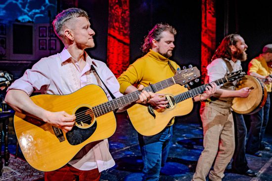 Two men in colourful trousers and shirts play acoustic guitars.