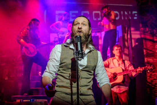 A male performer in a waistcoat singing into a microphone. There are various musicians behind him lit in red.