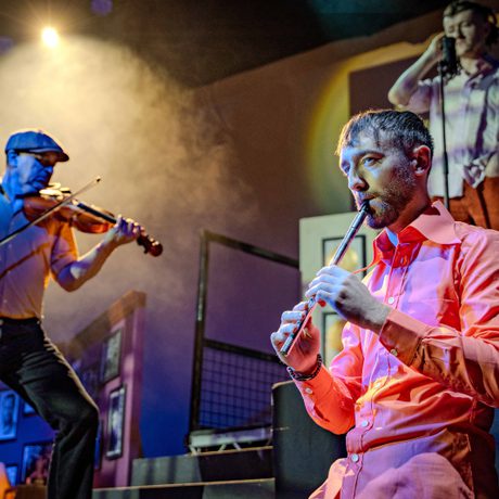 A male performer wearing a pink shirt playing a whistle. In the background another performer is playing the violin.