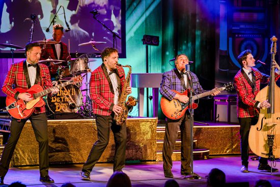 Four male musicians on a stage, playing guitar, double bass, and saxophone. Three men have a red tartan jacket, and one man has a purple tartan jacket.