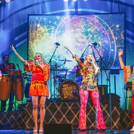 Four singers, two men and two women, wearing tie dye clothes and singing on a stage. They all have one arm in the air.