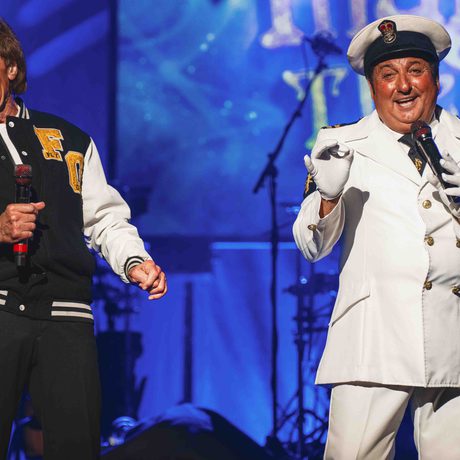 Two men on stage holding microphones and smiling. The man on the left is wearing a black jacket with white sleeves, and the man on the right is wearing a captain's uniform.