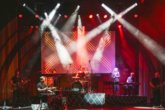 A stage with a band playing various instruments. The stage is lit red with multiple white spotlights.