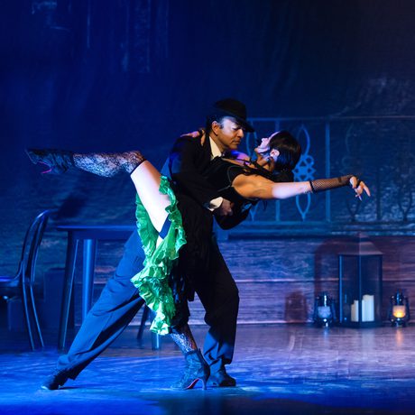 A man in a black suit and hat dances with a woman in a green and black dress.