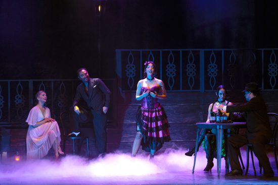 A stage filled with smoke and lit in purple. Three women, two in pink and one in green, and two men in black suits sit on chairs.