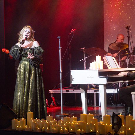 A white, blonde woman wearing a floor-length gold dress sings while a white man with white hair plays the piano. They are surrounded by candles.
