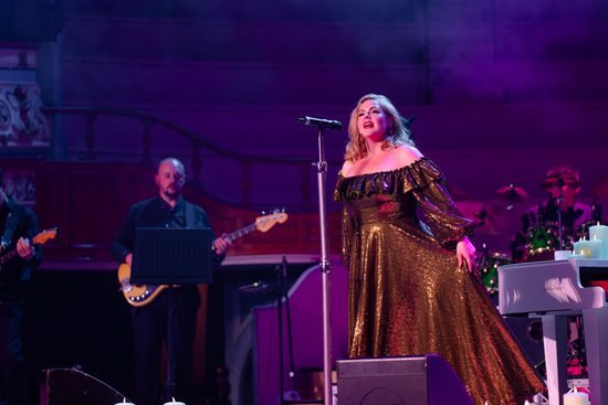 A white, blonde woman in a gold floor-length dress singing into a microphone.