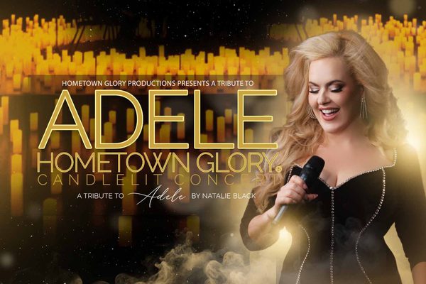 A blonde, white woman wearing a black dress holds a microphone and smiles. She is surrounded by lit candles.