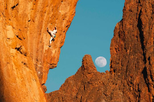 A climber on a cliff face of orange rock. The sky is vibrant blue and you can seen the moon.
